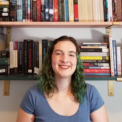 Headshot of Ashley Kempkes. She has medium long hair that is a mixture of brown and green. She is wearing a blue shirt. She is smiling. 