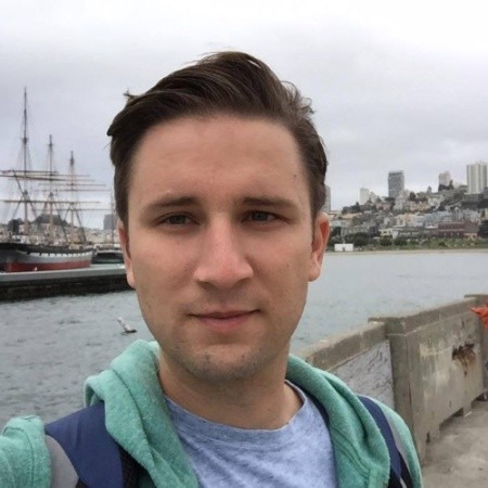 Picture of a man with short brown hair, wearing a grey shirt and a green jacket. 