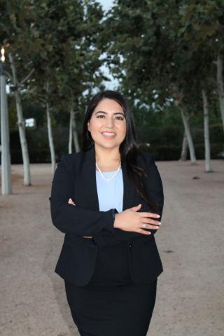 Picture of a woman with black hair, standing with her arms folded in a black jacket. 