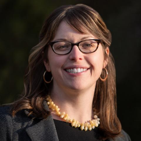 Picture of a women with long dark hair, wearing glasses, a dark top, and a yellow necklace. 