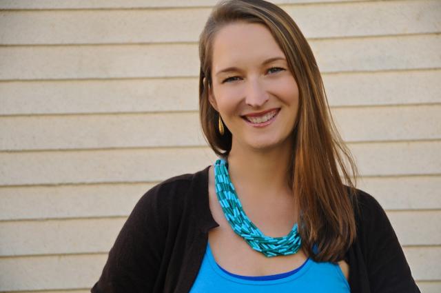 Picture of a woman with long, brown hair wearing a blue top, black sweater, and blue necklace. 