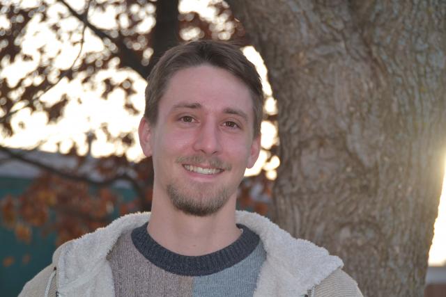 Picture of a man with short brown hair, wearing a grey shirt and a grey jacket. 