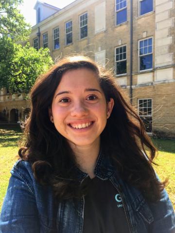 Headshot of Ana Belen. She has long brown hair. She is wearing a blue top and is standing in front of a UNT building on campus.  She is smiling. 