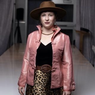 Picture of Kitty. She is standing, wearing a black top, pink jacket, leopard print skirt, and a brown hat. She is smiling. 