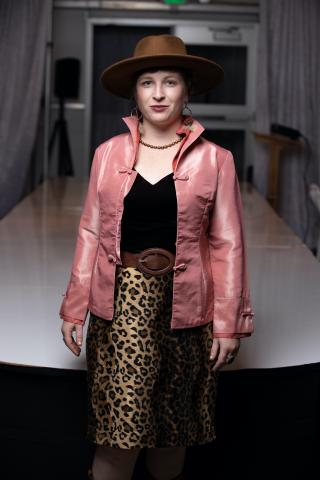 Picture of Kitty. She is standing, wearing a black top, pink jacket, leopard print skirt, and a brown hat. She is smiling.