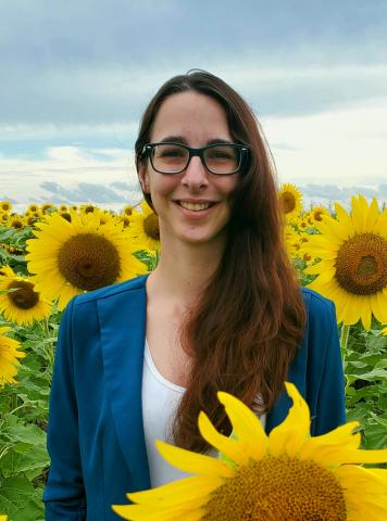 Headshot of Shannon.  She has long brown hair. She has on a blue shirt and is standing in a field of sunflowers. She is wearing glasses and is smiling. 