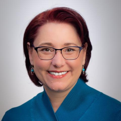 Picture of a woman with short red hair, wearing glasses and a blue top. 